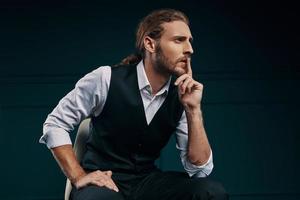 Thoughtful young man in elegant shirt looking away and keeping finger on lips while sitting against green background photo