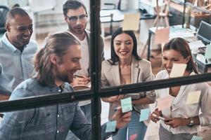 asegurando el éxito trabajando en equipo. vista superior de los jóvenes modernos con ropa informal inteligente usando notas adhesivas mientras están de pie detrás de la pared de vidrio en la sala de juntas foto