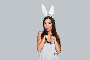 Funny bunny. Beautiful young Asian woman in bunny ears smiling while standing against grey background photo