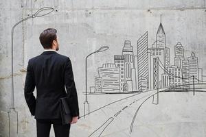 Moving forward. Rear view of young businessman standing against concrete wall with city sketch on it photo