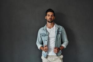 hombre perfecto. un joven apuesto mirando a la cámara y ajustando su chaqueta vaquera mientras se enfrenta a un fondo gris foto