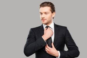 Making business look good. Confident young man in formalwear adjusting his necktie and looking away while standing against grey background photo