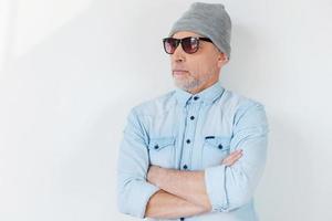 confiado en su estilo. hombre mayor de pelo gris confiado con gafas de sol y sombrero funky mirando hacia otro lado y manteniendo los brazos cruzados mientras está de pie contra el fondo blanco foto