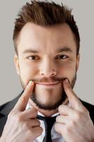 Making a fake smile. Portrait of young man in formalwear making a smile by his fingers while standing against grey background photo