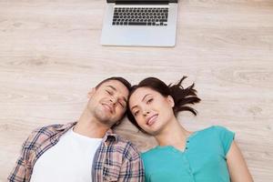 Love connecting. Top view of cheerful young couple lying down on the floor and looking at camera while laptop lying upon their heads photo