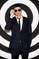 Charming macho. Handsome young man in full suit adjusting his sunglasses while standing against black and white background photo