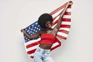 mujer africana joven feliz que lleva la bandera americana y sonriendo mientras está de pie contra el fondo gris foto