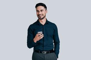 Receiving good news. Handsome young man holding smart phone and smiling while standing against grey background photo