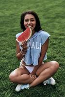Enjoying healthy snack.  Attractive young woman eating a slice of watermelon and smiling while sitting on the grass