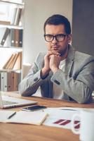 Making business every day. Handsome pensive young man in glasses looking at camera while sitting at his working place photo