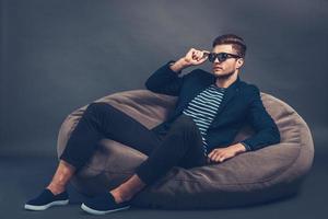 Handsome and stylish. Confident young handsome man adjusting his sunglasses and looking away while sitting on beanbag against grey background photo