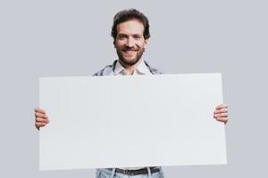 llevando espacio de copia. joven confiado vestido con ropa informal elegante que sostiene un rotafolio en blanco y sonríe mientras se enfrenta a un fondo gris foto