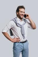 mejores decisiones. un joven hermoso con ropa elegante e informal hablando por teléfono y sonriendo mientras se enfrenta a un fondo gris foto