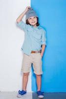 Checking up his style. Full length of handsome little boy adjusting his hat and looking up while standing against white and blue background photo