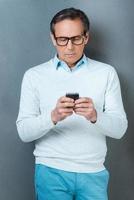 Using his phone for being in touch. Confident mature man holding mobile phone while standing against grey background photo