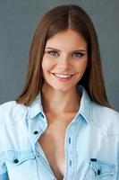 Sincere smile. Smiling young woman in shirt looking at camera while standing against grey background photo