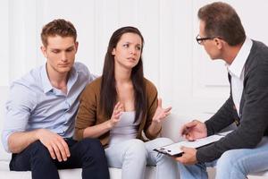 Asking for an expert advice. Frustrated young couple sitting on the couch while woman telling about their relationship problems to psychiatrist and gesturing photo