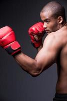 boxeador confiado. vista lateral de un joven africano sin camisa con guantes de boxeo haciendo ejercicio mientras se enfrenta a un fondo gris