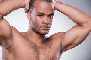 Strong and muscular man. Young African man holding hands behind head while standing isolated on grey background photo