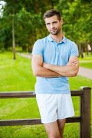 Charming handsome. Handsome young man keeping arms crossed and smiling while standing outdoors and leaning at the fence photo