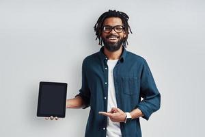Charming young African man in eyewear pointing copy space using digital tablet while standing against grey background photo