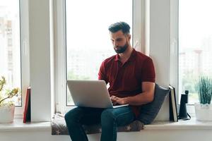 Concentrated young man in casual clothing using laptop while sitting on the window sill photo