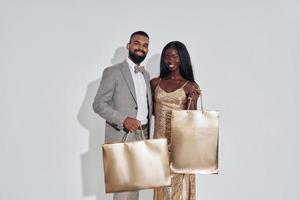 hermosa pareja africana que lleva bolsas de compras de color dorado y sonríe mientras está de pie contra el fondo gris foto