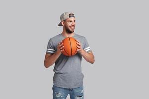 joven y lleno de energía. un apuesto joven sonriente que lleva una pelota de baloncesto y mira hacia otro lado mientras se enfrenta a un fondo gris foto