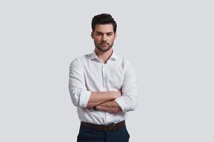 Pure perfection. Good looking young man looking at camera and keeping arms crossed while standing against grey background photo