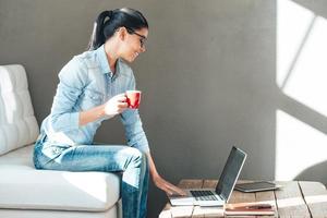 disfrutando de su día de trabajo. vista lateral de una hermosa joven alegre con gafas sosteniendo una taza de café y trabajando en una laptop con una sonrisa mientras se sienta en el sofá de la oficina