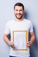 Copy space in frame. Handsome young man in T-shirt holding a picture frame while standing against grey background photo