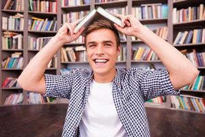 Feeling playful. Cheerful young man carrying book on head and smiling while sitting against bookshelf photo