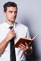 Waiting for inspiration. Thoughtful young man in shirt and tie holding note pad and touching his chin with pen while standing against grey background photo