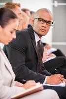 Businessman at the conference. Side view of business people sitting in a row and writing something in their note pads while confident African man in eyewear looking at camera photo