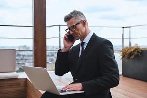Confident mature man working on laptop and using mobile phone while standing on the rooftop terrace photo