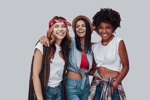 tres atractivas mujeres jóvenes con estilo mirando a la cámara y sonriendo mientras están de pie contra el fondo gris foto