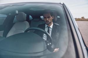 Hurrying to get things done. Handsome young man in full suit looking straight while driving a car photo