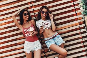 regalo de verano perfecto. dos atractivas mujeres jóvenes sonriendo y sosteniendo un helado mientras se apoyan en la pared de madera al aire libre foto