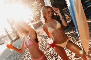verano lleno de alegría. dos atractivas mujeres jóvenes sonriendo y disfrutando de cócteles mientras se divierten en la playa foto