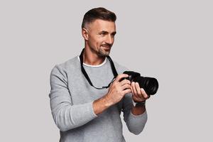 Handsome journalist. Good looking young man holding digital camera and smiling while standing against grey background photo