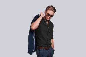 Charming young man. Good looking young man in eyewear carrying jacket on shoulder while standing against grey background photo