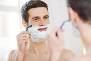 Morning routine. Handsome shirtless young man shaving his face and smiling while standing in front of the mirror photo