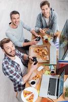 les gusta lo que hacen. vista superior de tres jóvenes jugando juegos de computadora y comiendo pizza mientras están sentados en el escritorio foto