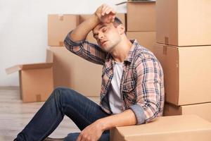 Tired of moving boxes. Tired young man sitting on the floor and leaning at the carton box stack while more cardboard boxes laying on the background photo