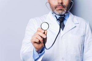 Medical exam. Close-up of mature grey hair doctor examining you with stethoscope while standing against grey background photo