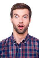 Shocking news. Portrait of surprised young man in casual shirt keeping mouth open while staring at camera while standing isolated on white photo