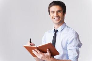 hombre de negocios con bloc de notas. un joven apuesto con camisa y corbata sosteniendo un bloc de notas y sonriendo mientras se enfrenta a un fondo gris foto