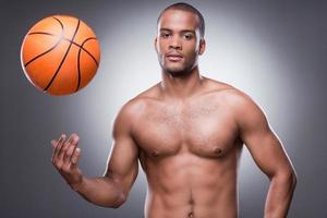 esperando un juego. joven africano sin camisa lanzando una pelota de baloncesto y mirando a la cámara mientras se enfrenta a un fondo gris foto