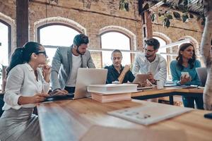 Group of young modern people in smart casual wear communicating and using modern technologies while working in the office photo