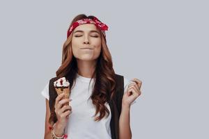 Attractive stylish young woman eating ice cream and smiling while standing against grey background photo